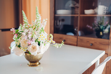 Fresh flower arrangement made in brass vase put on dining table decorating kitchen interior. White beige roses, digitalis blooms