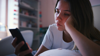 Depressed Teenage Girl Lying On Bed At Home Looking At Mobile Phone