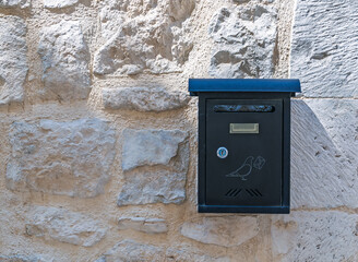 Modern black mailbox with a decorative bird graphic mounted on a rustic ancient stone wall in bright sunlight.Contrast of old and new