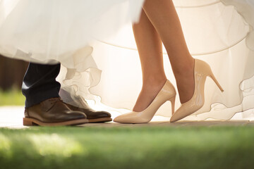 Close-up of a bride in high heels and groom in classic shoes under a white wedding dress.