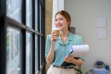 Young Asia businesswoman drinking coffee and holding documents while looking out the window in modern office