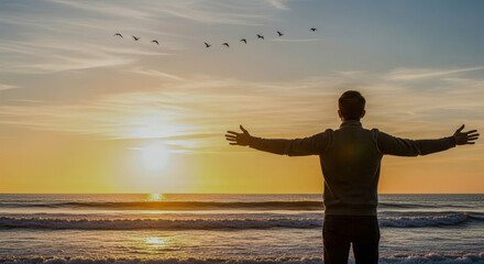 Rear view of a man with arms outstretched at the beach during sunset, birds flying across the sky. Freedom and wellbeing.
