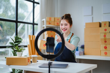 Happy asian seller preparing packages for delivery, using smartphone and ring lamp, recording video for social media