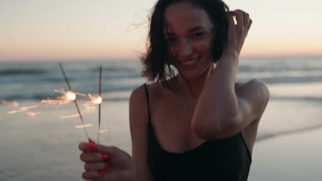 Carefree young woman laughs and smiles as she plays with sparklers on the beach at sunset. The gorgeous golden hour light bathes the scene in warmth.