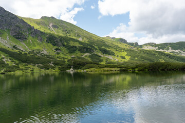 Serene mountain lake reflecting green hills and cloudy sky in pristine alpine landscape with calm crystal clear water. Concept of natural beauty, tranquility and wilderness conservation.