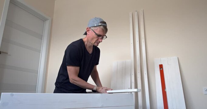 Man assembling white wooden shelving unit at home, measuring and drilling parts with precision, DIY furniture construction process. Men collects a wooden shelf at home with his own hands