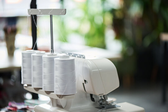 Industrial overlock sewing machine standing on table with multiple large thread spools attached, threads running through guides, blurred workspace background visible