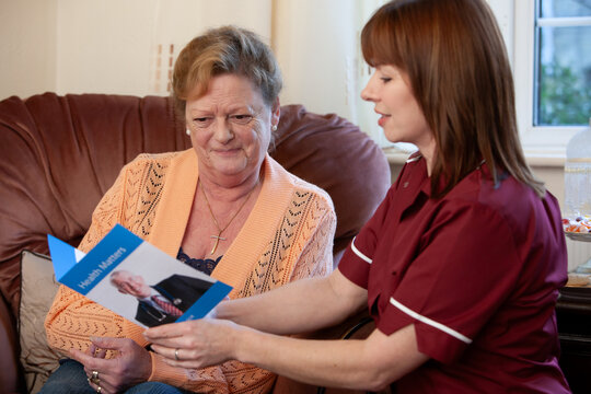 A care worker shows an elderly woman a health leaflet during a home visit as they discuss the information provided.