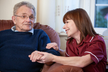 An elderly man visited at home by his friendly care worker who holds his hand listening intently with a smile while he talks to her.