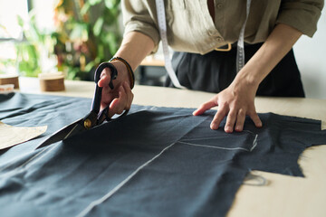 Caucasian young adult woman cutting fabric with large scissors on worktable, hands visible holding textile, measuring tape draped around neck, preparing garment pattern