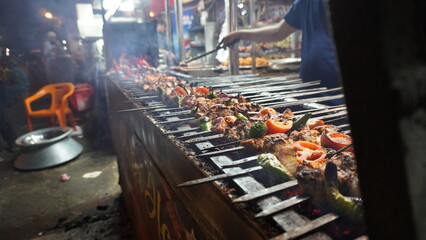 Long grill with kebab skewers cooking, smoke rising, outdoor setting at night, with dark shadowy areas.