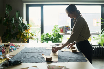Young adult Caucasian woman working with fabric and paper patterns on large table, using pattern weights and measuring tape, focused on preparing textile pieces in bright studio