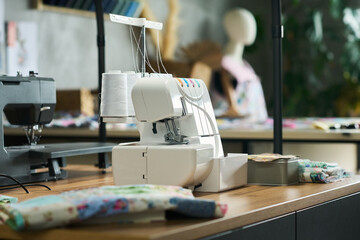 Serger sewing machine standing on wooden table surrounded by colorful fabric pieces in modern workspace, blurred mannequin and sewing supplies visible in background