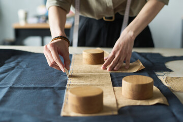 Young adult Caucasian woman marking fabric with tailor chalk while holding paper sewing pattern and pattern weights on worktable, focusing on precise garment construction process