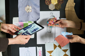 Caucasian woman holding tablet displaying fashion sketches while another Caucasian woman presenting fabric swatches, collaborating on textile design project in creative workspace
