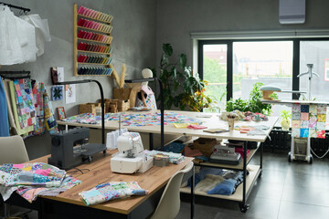 Sewing studio showing large worktable covered with colorful fabric pieces, sewing machines, spools of thread on wall rack, patchwork quilts, bright window with plants, no people visible