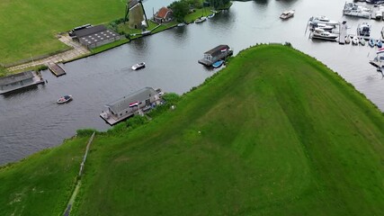 A calm aerial scene of floating houses on a river, surrounded by vast green meadows and intersecting waterways with Dutch flags gently waving in the breeze under a cloudy sky. A classic Dutch windmill