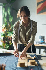 Young adult Caucasian woman working with sewing patterns on fabric, measuring and cutting material in workshop, tape measure around neck, focused on garment making process