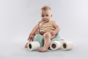 Cute little baby sitting on potty and rolls of toilet paper against light grey background
