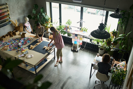 Three Caucasian adults working in sewing studio, two standing and cutting fabric on large table while one sitting at sewing machine, surrounded by colorful textiles and plants - Powered by Adobe