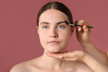 Fototapeta premium Cosmetologist brushing young woman's eyebrow with spoolie on pink background, closeup