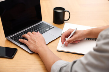 Fototapeta premium Copywriter using laptop and writing in notebook at wooden table indoors, closeup