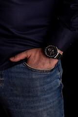 Close-up of man hand casually resting in denim pocket, highlighting fashionable wristwatch with intricate metallic details, set against dark background for contrast and emphasis. Men's watch