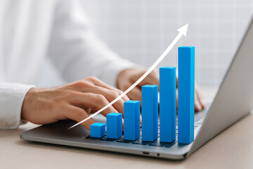Close up of hands typing on a laptop with a rising blue bar graph and upward arrow overlay indicating financial growth