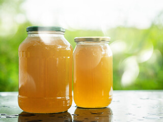 Two glass jars of golden bee honey are on a wet table, green grass field and sky in the background. Fine organic product concept. Agriculture and food industry. Light and airy feel and mood.