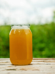 Glass jar of golden bee honey is on a wooden table, green grass field and sky in the background. Fine organic nature product concept. Agriculture and food industry.