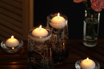 Burning candles, spa stones and flowers in glasses of water on table against black background, closeup