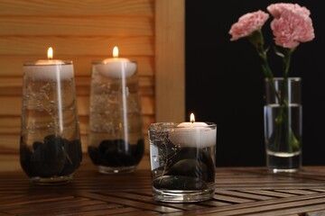 Burning candles, spa stones and flowers in glasses of water on wooden table against black background
