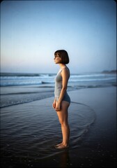 A serene side view of a young woman with short brown hair and glasses standing on a beach, lost in thought as gentle waves lap at her feet during the tranquil twilight hour .