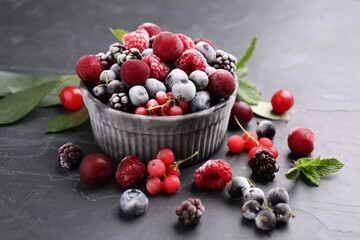 Mix of ripe frozen berries, leaves and bowl on dark textured table, closeup