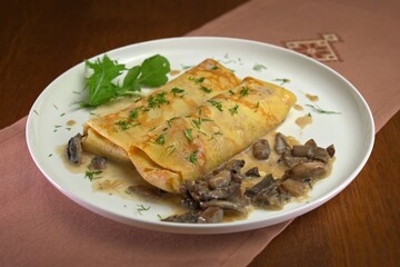 Savory crepes filled with mushrooms and onions, topped with creamy mushroom sauce and garnished with fresh dill on a white plate.