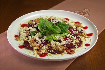 Fresh beetroot carpaccio topped with crumbled feta, walnuts, mixed greens, and berry sauce on a white plate.