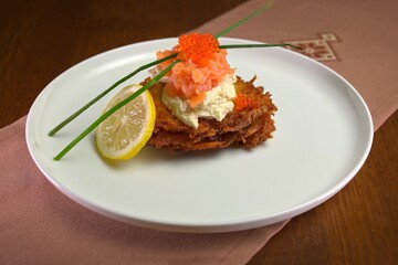Golden potato pancakes topped with cream cheese, diced smoked salmon, and red caviar, garnished with chives and lemon slice on a white plate.