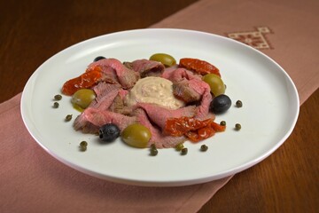 Vitello tonnato-style dish with thin slices of rare beef, tuna sauce, olives, sun-dried tomatoes, and capers on a white plate.
