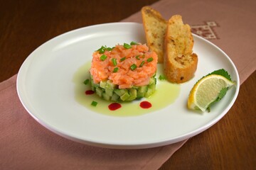 Elegant salmon tartare served on a bed of diced cucumber and avocado, garnished with chives, red sauce drops, lemon slice, and toasted baguette slices.