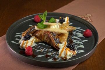 Triangular folded crepes served with whipped cream, caramel drizzle, blackberries, raspberries, wafer sticks, and mint, presented on a dark plate with artistic sauce decoration.