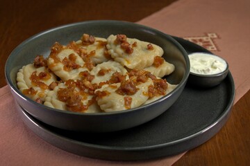 Traditional dumplings topped with crispy fried onions and bacon, served with a side of sour cream in a dark ceramic bowl.