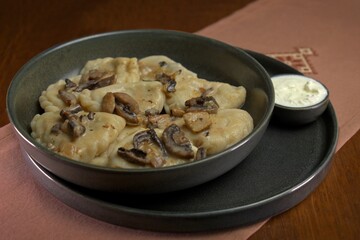 Dumplings topped with saut&eacute;ed mushrooms and onions, served with a side of sour cream in a rustic ceramic bowl.