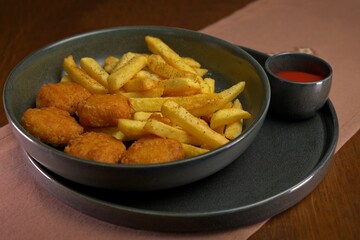 Crispy chicken nuggets with seasoned French fries served in a dark bowl, accompanied by a small cup of red dipping sauce.