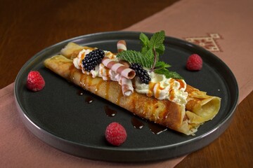 Rolled crepe dessert topped with whipped cream, caramel sauce, blackberries, mint leaves, and wafer rolls, served on a black plate with raspberries.