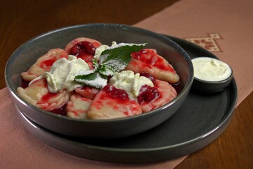 Sweet dumplings filled with cherries, served with cream and powdered sugar, garnished with fresh mint in a black ceramic bowl.