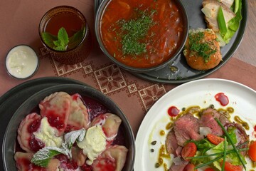 Top view of traditional Eastern European meal including borscht, raspberry dumplings, roast beef salad, bread, and iced tea on embroidered linen.