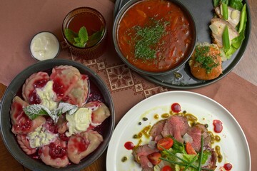 Top view of traditional Eastern European meal including borscht, raspberry dumplings, roast beef salad, bread, and iced tea on embroidered linen.