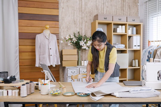 Asian female fashion designer sketching lines on large white paper using pencil before cutting fabric in home studio workspace with tools threads and garments representing creative startup business