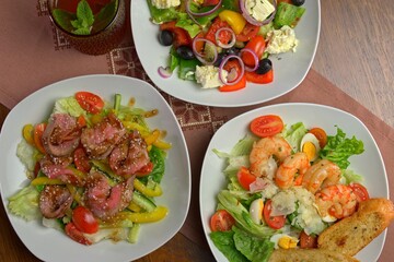 Top view of three fresh salads with grilled beef, shrimp, and Greek-style ingredients, served on white plates with a refreshing drink in a glass on a traditional embroidered napkin.