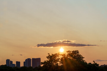 The sun sets below a glowing cloud over a distant city skyline, casting warm light above tree silhouettes, Scarborough, Ontario, Canada